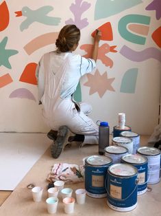 a woman painting a wall with paint cans on the floor next to her and several other paints