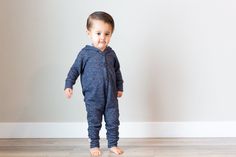 a toddler standing in front of a wall wearing a blue onesie and smiling