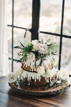 a wedding cake with white flowers and greenery on a wooden table in front of a window