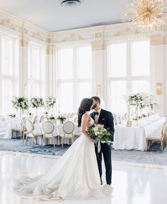 a bride and groom standing in front of a banquet room with white tables, chandeliers and floral centerpieces