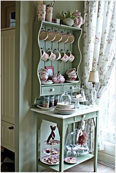 a green china cabinet with plates and cups on it's shelf next to a window