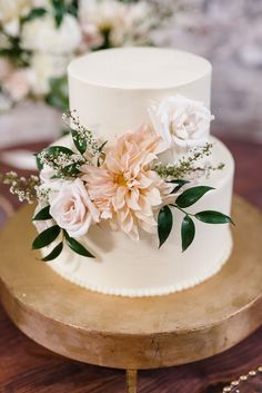 a white wedding cake with pink flowers and greenery on the top is sitting on a wooden stand