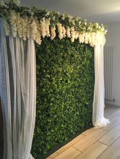 an artificial green wall with white flowers and sheer drapes on the top, in front of a wooden floor