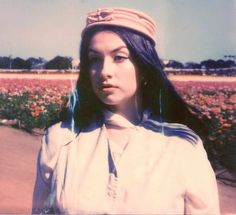 a woman with long black hair wearing a hat in a flowered field and looking off to the side