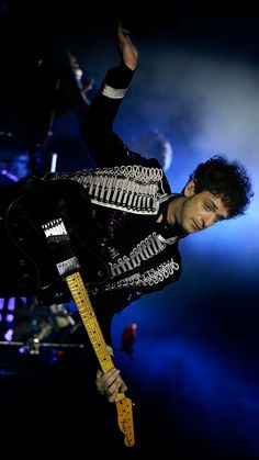 a man holding a guitar in his right hand and wearing a black jacket with silver details on it