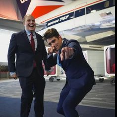 two men in suits pointing at the camera while standing next to an airplane with its door open