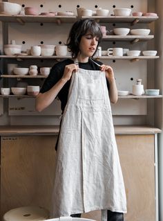 a woman standing in front of a shelf with bowls and plates on it, holding a white apron over her shoulder