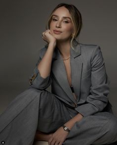 a woman sitting on top of a stool wearing a gray suit and gold jewelry, with her hand under her chin