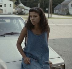 a woman standing next to a parked car on the side of the road with her hands in her pockets