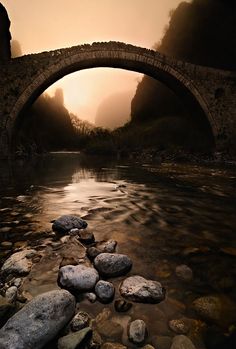 a stone bridge over a river with rocks in the foreground and mountains in the background