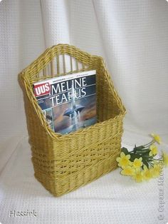 a magazine in a wicker basket next to yellow flowers on a white cloth background