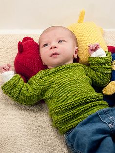 a baby laying on the floor wearing a green sweater and holding two red stuffed animals