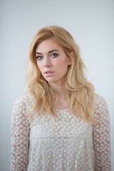 a woman with long blonde hair wearing a white top and looking at the camera while standing in front of a wall