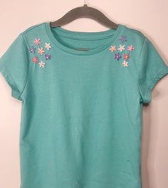 a green t - shirt with flowers on it hanging from a hanger in front of a white wall