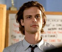 a man with long hair wearing a shirt and tie in front of a whiteboard