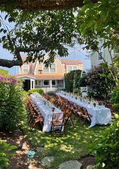 a long table set up in the yard for an outdoor dinner party with white linens and wooden chairs