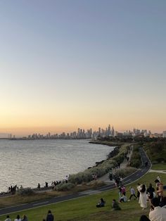 many people are sitting on the grass by the water and looking at the city in the distance