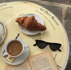 an open book, coffee cup and croissant on a table with reading glasses