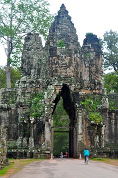 two people are riding bicycles in front of the entrance to an ancient building that is surrounded by trees