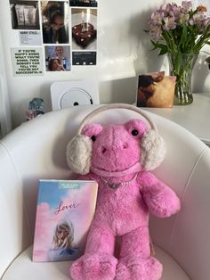 a pink teddy bear with headphones sitting on a chair next to a book and flowers