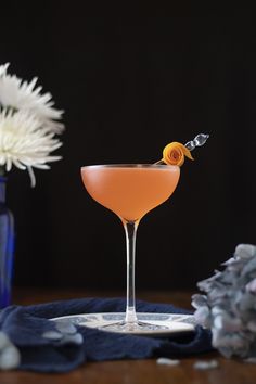 a drink in a coupe glass with an orange garnish on the rim next to flowers