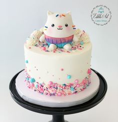 a white cake with sprinkles and a cat sitting on top of it