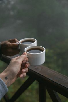 two people holding cups of coffee in their hands on a wooden deck overlooking the forest