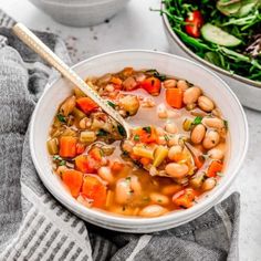 a white bowl filled with bean and vegetable soup on top of a gray towel next to a salad