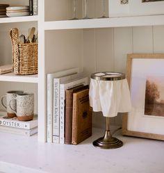 a white book shelf with books and cups on it, along with two framed pictures