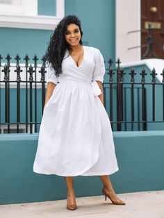 a woman wearing a white dress standing in front of a blue wall and black fence