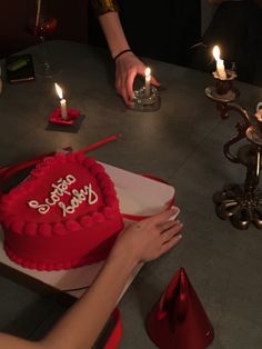 a red heart shaped cake sitting on top of a table next to two people holding lit candles