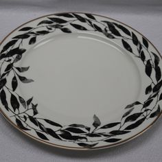 a white plate with black leaves on it and gold trim around the edge is sitting on a gray tablecloth