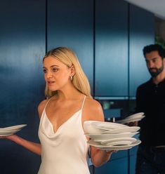 a woman in a white dress holding plates and looking at the camera while another man stands behind her