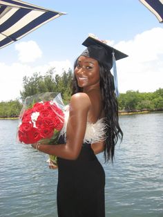 a woman in a graduation cap and gown is holding a bouquet of roses by the water