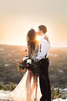 a bride and groom kissing on top of a mountain with the sun setting behind them