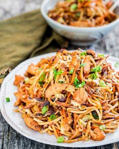 a white plate filled with noodles and meat on top of a wooden table next to a bowl