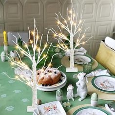 the table is set with plates, dishes and trees decorated with white lights on them