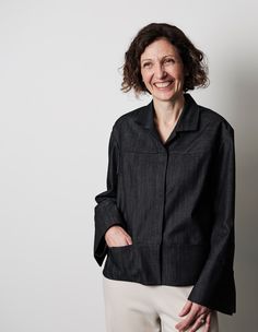 a woman standing in front of a white wall wearing a black shirt and beige pants