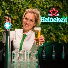 a woman is holding up a beer in front of many glasses on a bar with greenery behind her