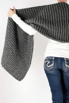 a woman is holding up a gray knitted shawl over her shoulder and looking down at the ground