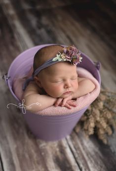 a newborn baby is sleeping in a purple bucket on a wooden floor wearing a flower headband