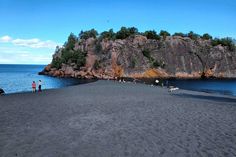 people are standing on the black sand beach near an island with some trees in it