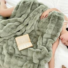 a woman laying in bed under a fluffy green blanket with an open book on it