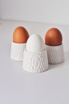 three eggs are sitting in white bowls on a table, one is brown and the other has an egg shell