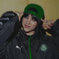 a woman with black hair wearing a green hat and jacket, smiling at the camera