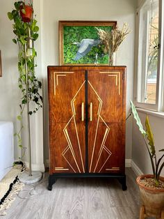 an art deco cabinet in the corner of a room with potted plants and a painting on the wall