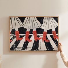 a woman is holding up a painting with red boots on it in front of a white wall