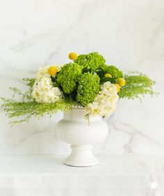 a white vase filled with lots of green and yellow flowers on top of a table