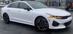 the front end of a white car parked in a parking lot next to other cars