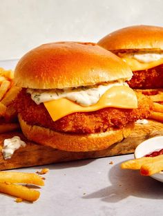 two chicken sandwiches with cheese and french fries on a cutting board next to some ketchup